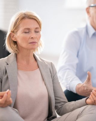 Aged blonde female in suit and her colleagues meditating at break while sitting in pose of lotus